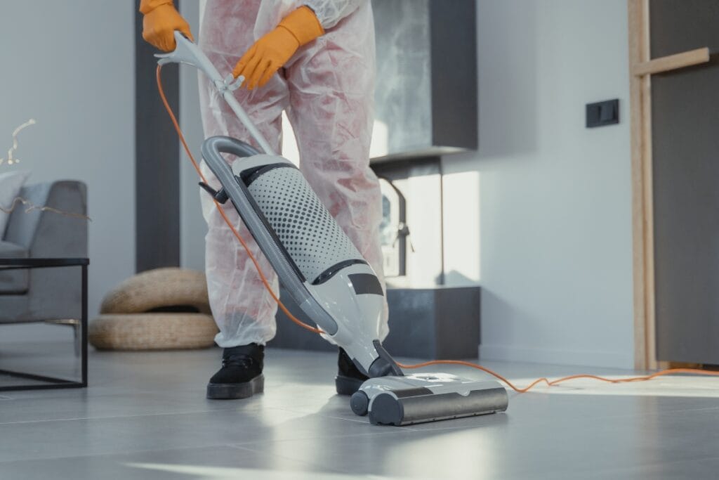 A person wearing protective clothing and orange gloves vacuums a light-colored tile floor in a modern, well-lit room, ensuring thorough cleaning for allergy sufferers.
