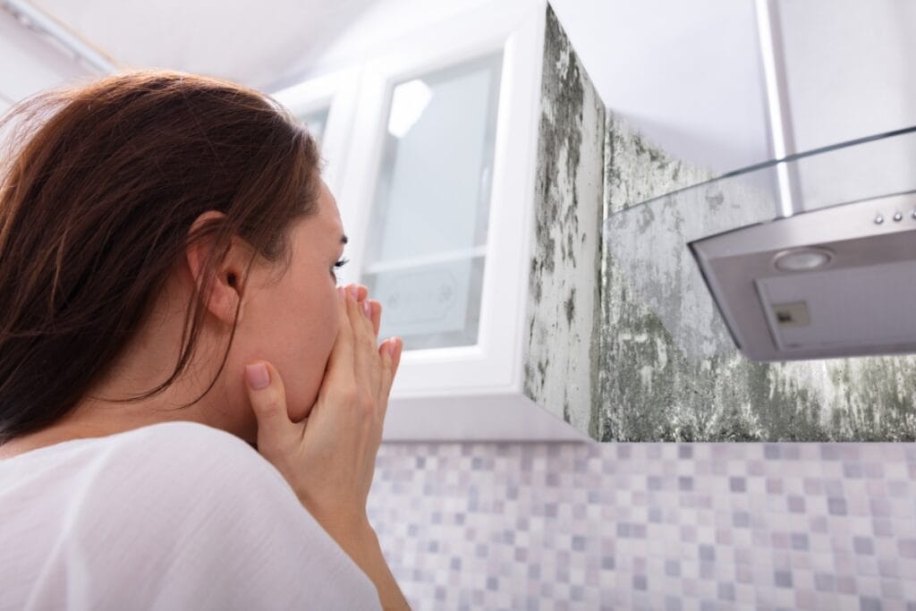 A woman covers her mouth while looking at mold growing on the kitchen wall near a range hood and cabinets, highlighting the importance of cleaning for allergy sufferers.