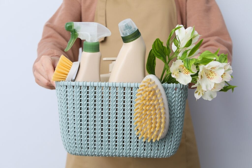 A person holding a blue woven basket containing cleaning spray bottles, scrub brushes, and white flowers—perfect for cleaning for allergy sufferers.