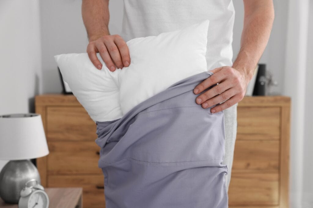 A person is putting a white pillow into a purple pillowcase in a bedroom with a wooden dresser and bedside lamp, taking steps toward cleaning for allergy sufferers.