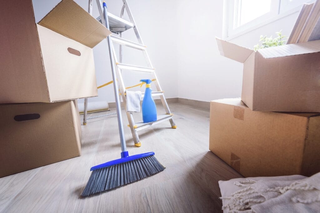 A broom, cleaning spray, and open cardboard boxes are arranged in a bright room with a stepladder, capturing the organized energy of move-out cleaning services or relocation tasks.