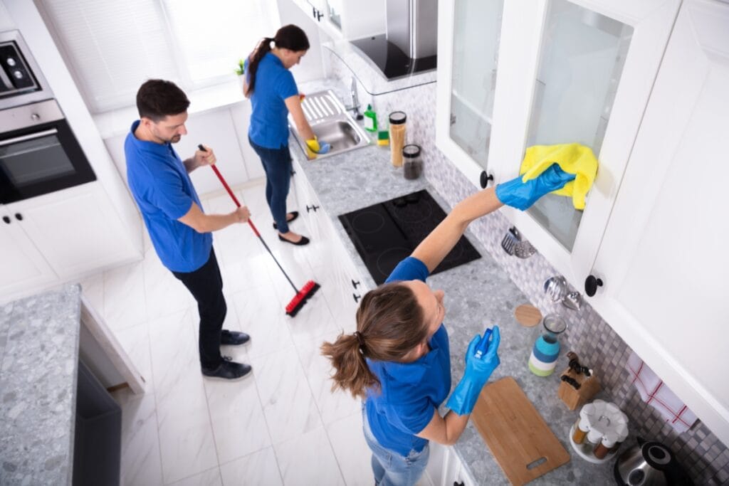 Three people in blue shirts provide move-out cleaning services in a modern white kitchen; one wipes a cabinet, another sweeps the floor, and a third washes dishes at the sink.