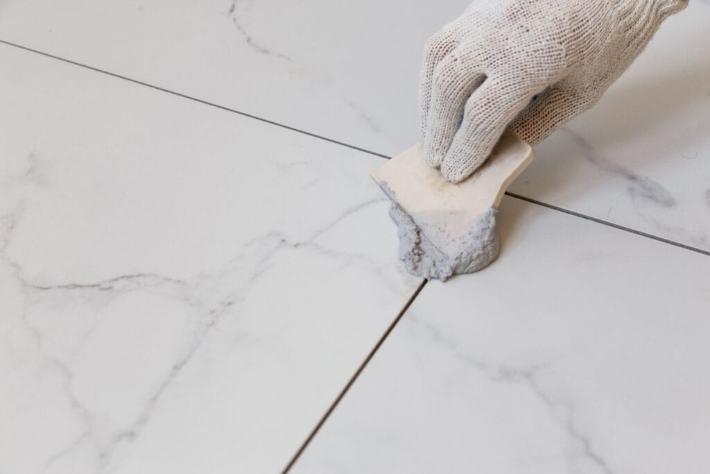 A gloved hand uses a tool to apply grout between white marble tiles, ensuring a flawless finish ideal for tile and grout cleaning.