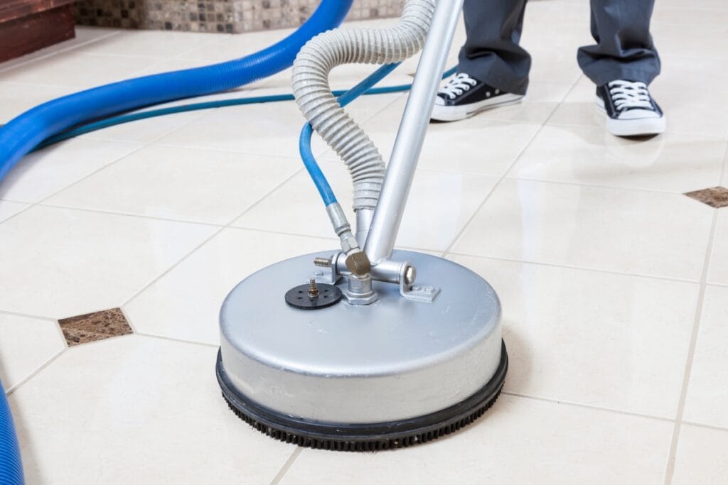 A person performing tile and grout cleaning with a professional machine on a tiled floor; blue hoses and part of the person’s legs are visible.