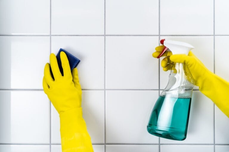 Person wearing yellow gloves cleaning white tiled wall with a blue sponge in one hand and a spray bottle with blue liquid in the other, demonstrating effective tile and grout cleaning techniques.