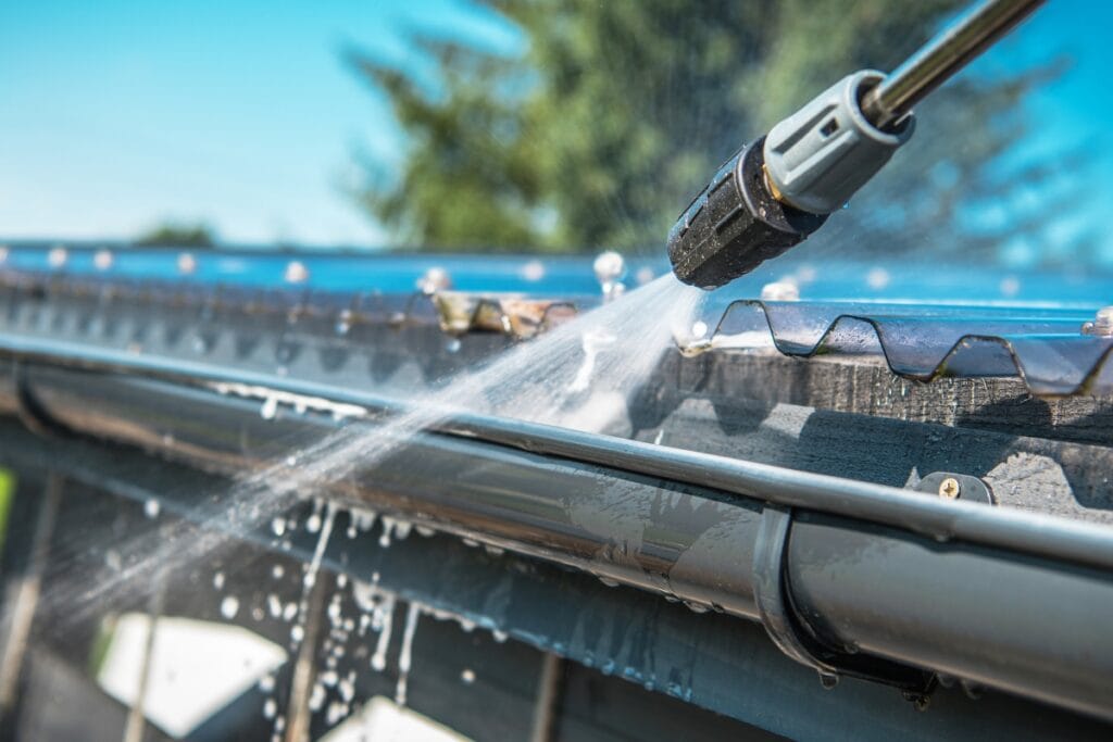 A pressure washer sprays water onto a house gutter, cleaning debris from the rooftop edge on a sunny day&mdash;showing how regular pressure washing keeps your home&rsquo;s exterior spotless.