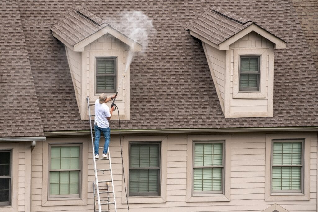 A person stands on a ladder using a power washer to clean the upper exterior of a house with multiple windows and shingled roofing, demonstrating the benefits of regular pressure washing for home maintenance.