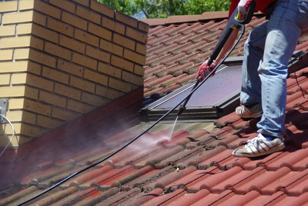A person uses a pressure washer to clean red roof tiles next to a brick chimney and a skylight, demonstrating how regular pressure washing keeps your roof looking fresh and well-maintained.