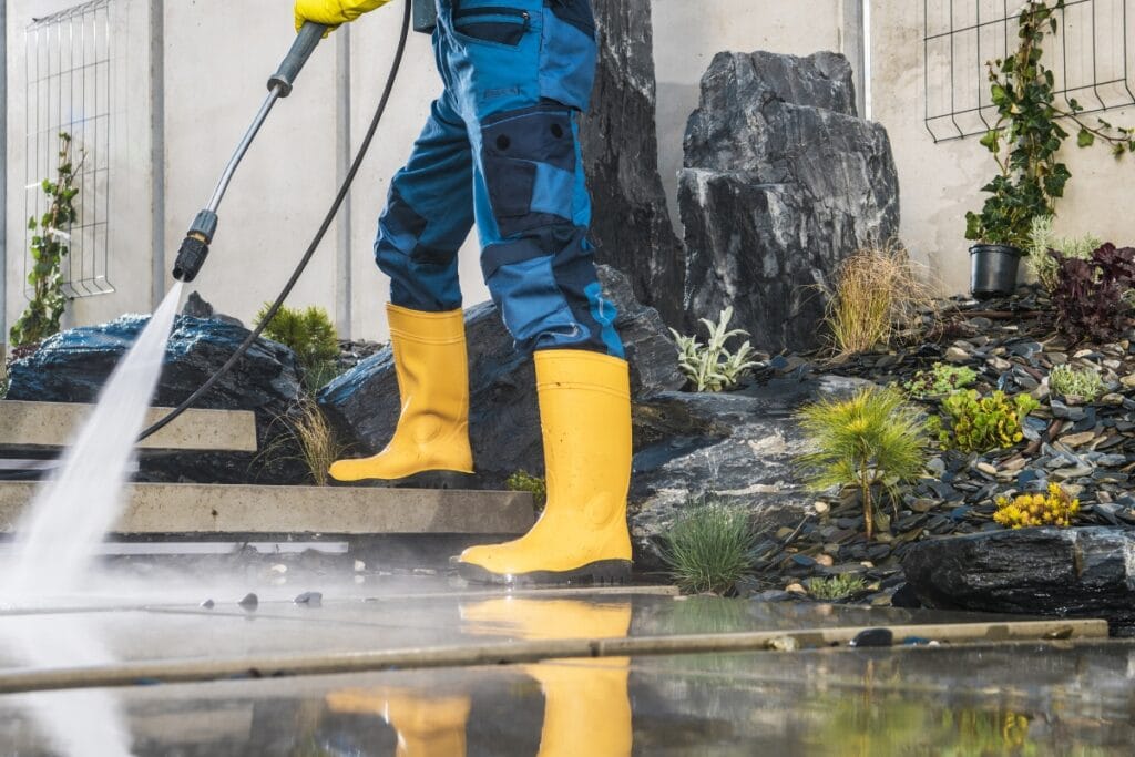 A person in yellow boots and blue coveralls uses a pressure washer to clean an outdoor patio area near a rock and plant garden, showing the benefits of regular pressure washing for maintaining spotless outdoor spaces.