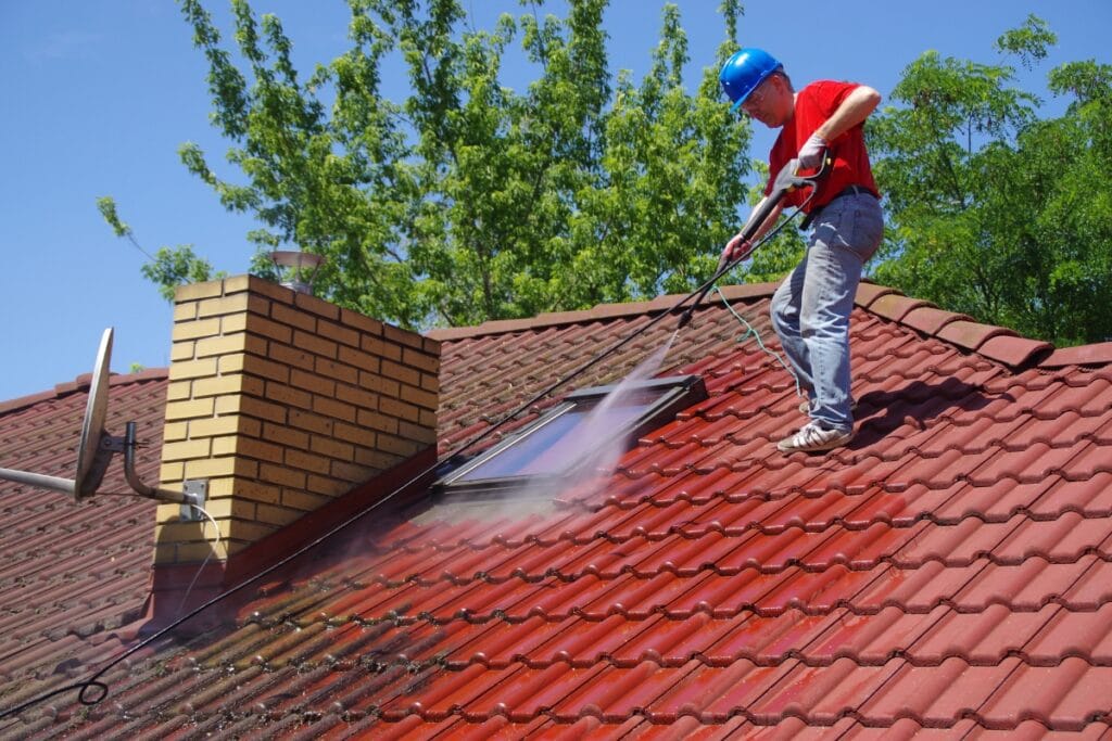 Man wearing a blue hard hat uses a pressure washer to clean a red tiled roof next to a chimney and skylight, highlighting the benefits of regular pressure washing.