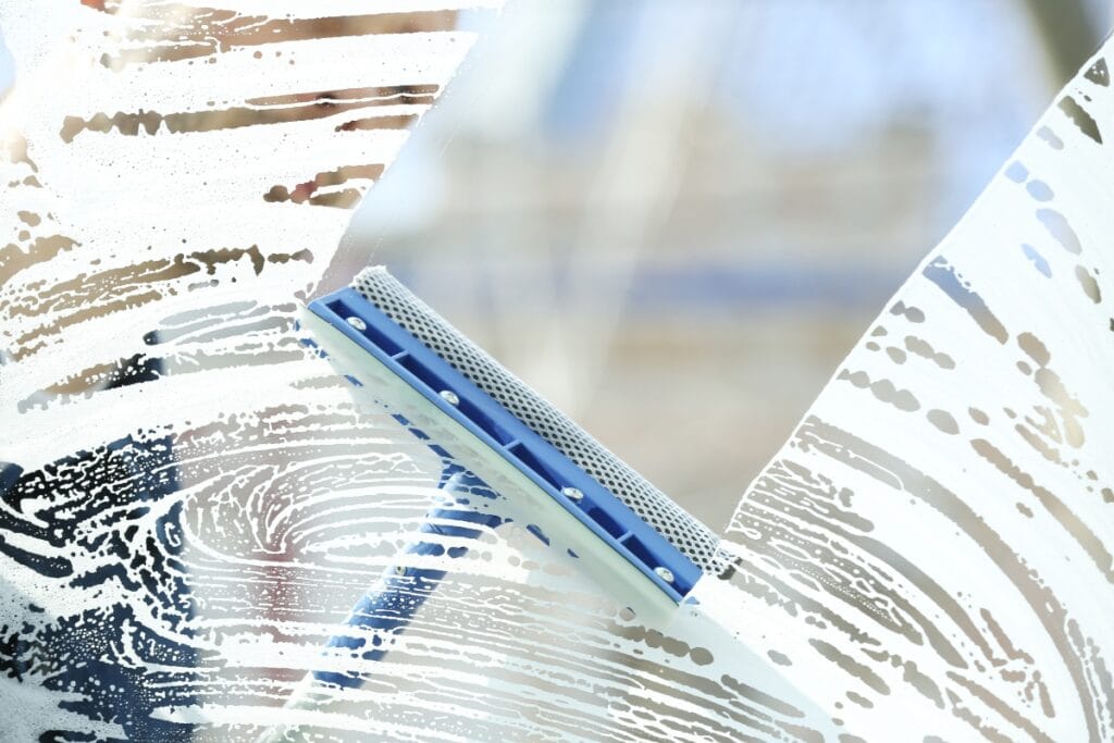 A close-up of a blue window squeegee cleaning soapy water from a glass surface, revealing a clear area amidst swirling soap suds—perfect technique that helps you avoid common window cleaning mistakes.