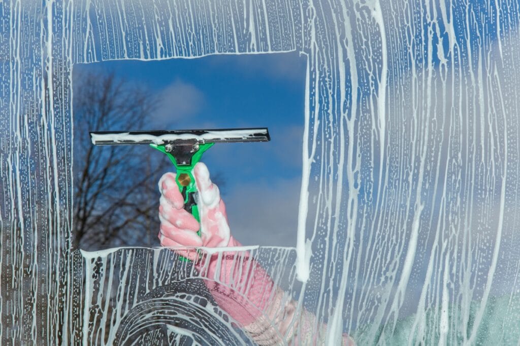 A person wearing a pink glove uses a squeegee to clean soap from a window, avoiding common window cleaning mistakes and revealing a clear view of the sky and trees outside.
