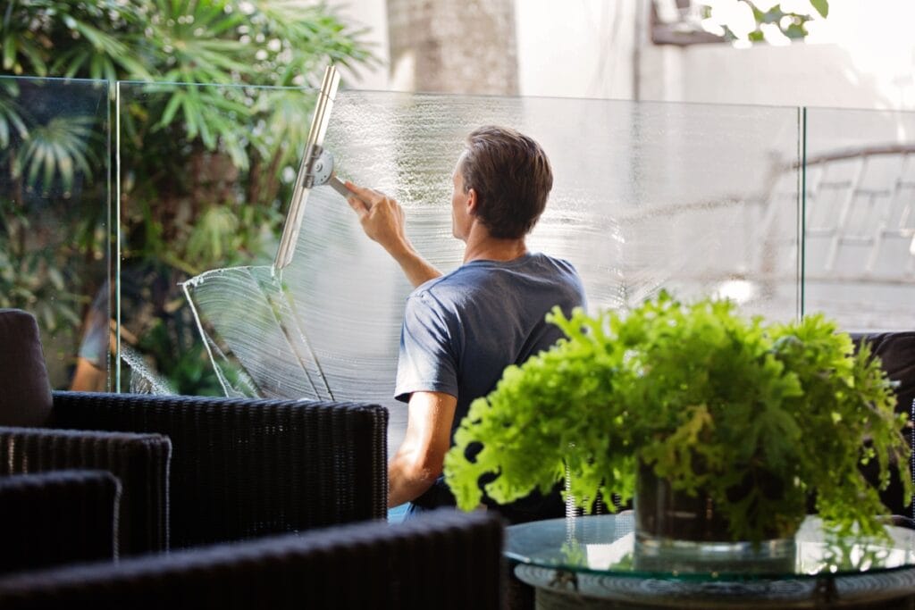 A person in a blue shirt carefully cleans a large glass window with a squeegee, avoiding common window cleaning mistakes, surrounded by wicker furniture and green plants.