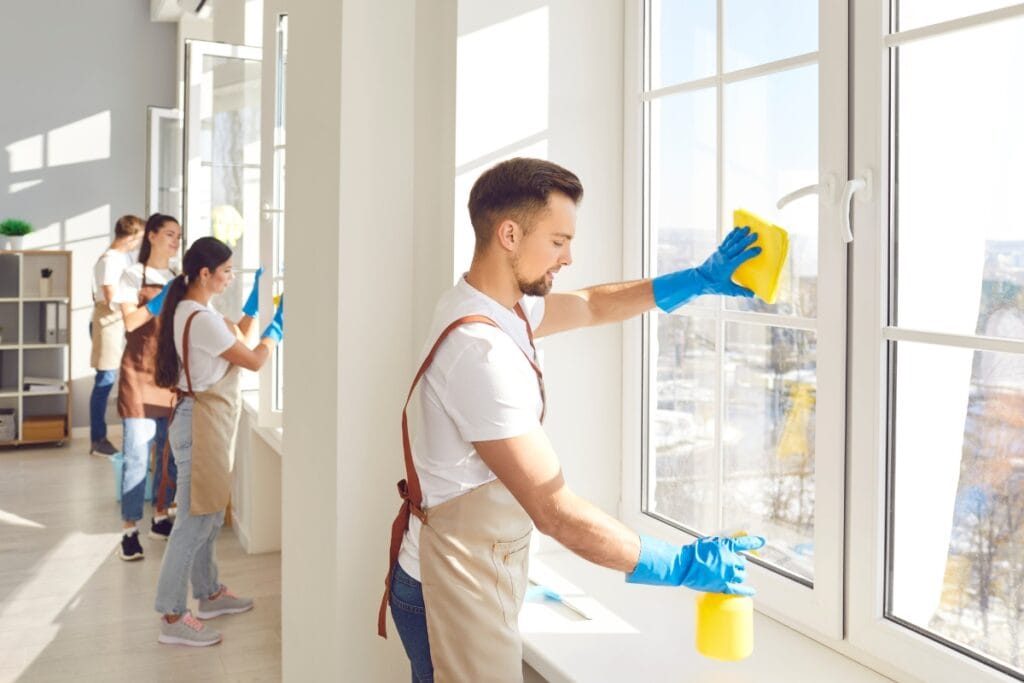 A group of people wearing aprons and gloves clean large windows inside a bright room, taking care to avoid common window cleaning mistakes.