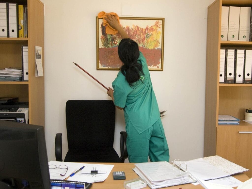 A person in green scrubs dusts the top of a framed picture on a wall in an office, showcasing the Best Office Cleaning practices while standing behind a black chair and desk with papers and office supplies.