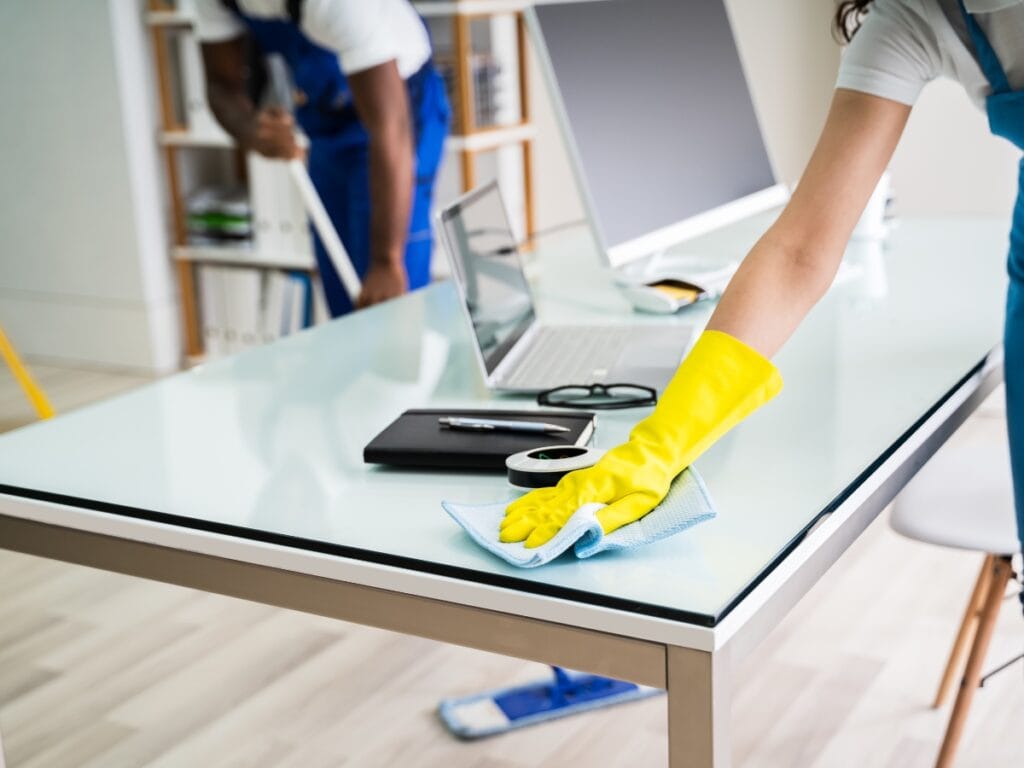 Two people providing Best Office Cleaning services: one wipes a desk with a cloth while wearing yellow gloves, and the other mops the floor in the background.