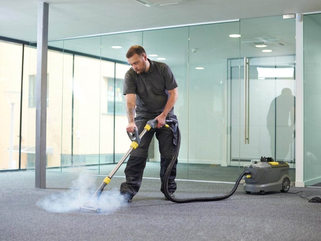 A man uses a steam cleaning machine to clean the carpet in a modern office space with glass walls, providing the best office cleaning for a fresh and professional environment.