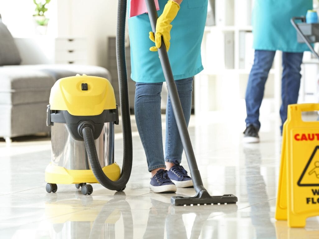 A person wearing gloves and an apron uses a vacuum cleaner to clean a shiny floor; a caution wet floor sign is visible nearby, showcasing the Best Office Cleaning practices.