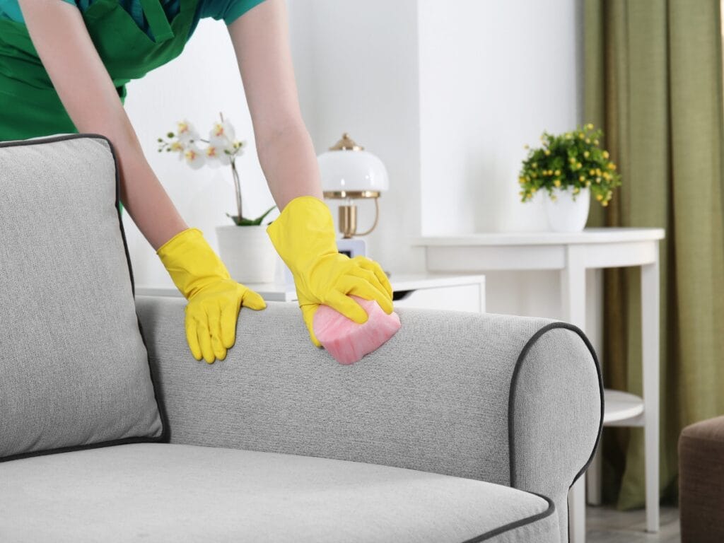A person wearing yellow rubber gloves performs upholstery cleaning, using a pink sponge to clean a light gray sofa in a living room with white furniture and green plants.