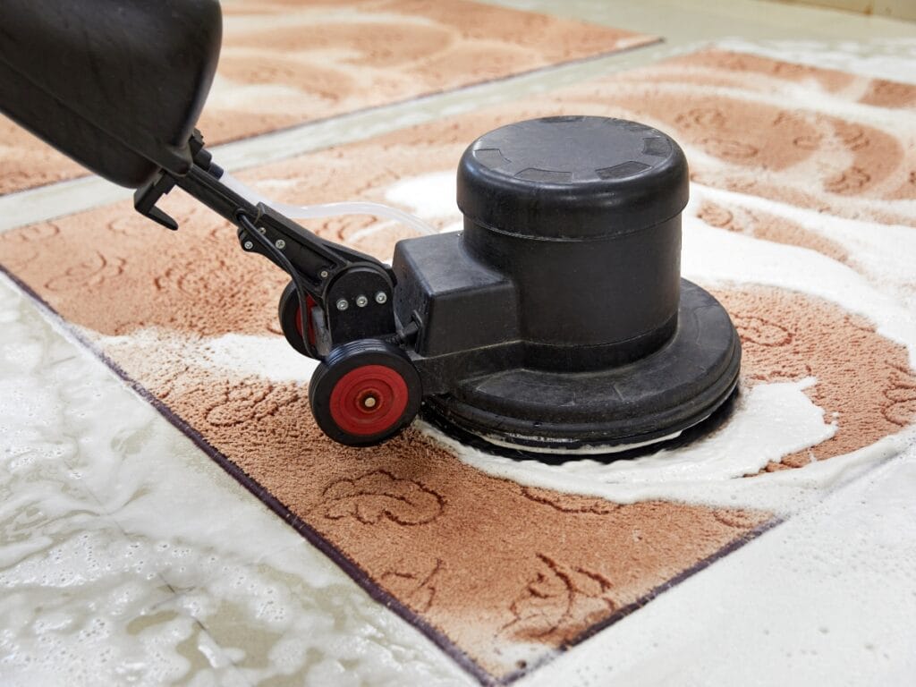 A floor cleaning machine scrubs a carpet with soap and water, creating foamy patterns on the surface to help extend carpet lifespan.