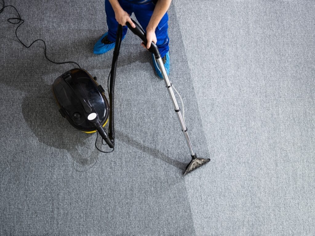 A person cleaning a gray carpet with a vacuum cleaner, showing a clear division between the cleaned and uncleaned sections to help extend carpet lifespan.