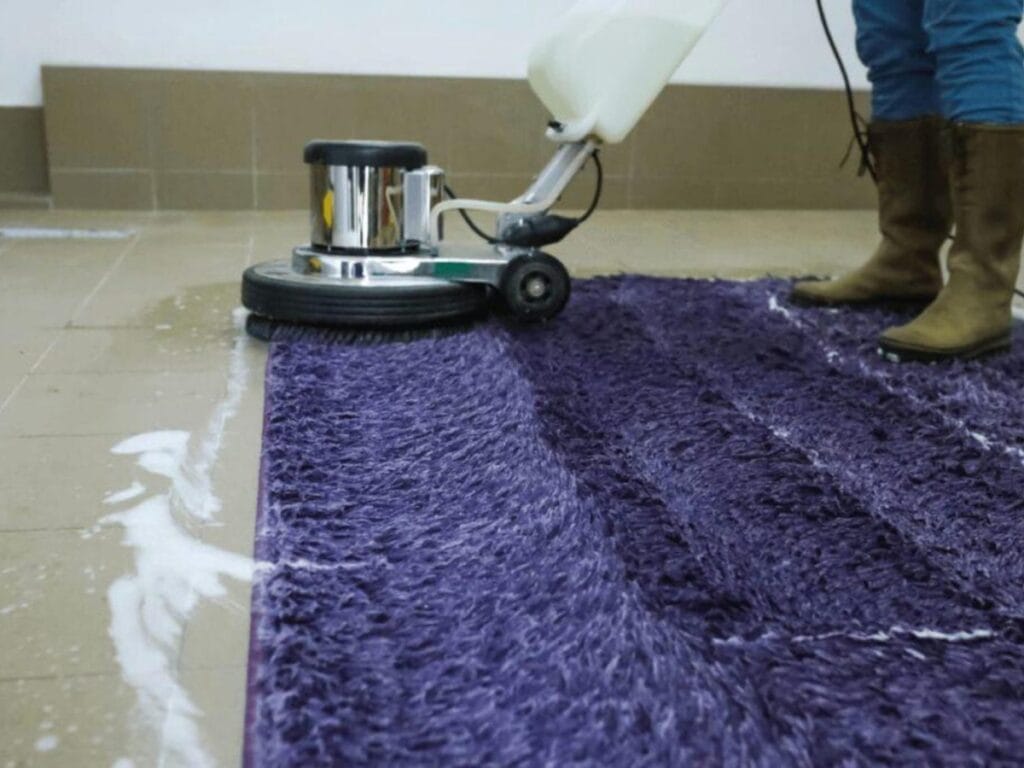 A person uses a floor cleaning machine to scrub a purple carpet, creating a clean path with soap suds on the tiled floor&mdash;helping to extend carpet lifespan.