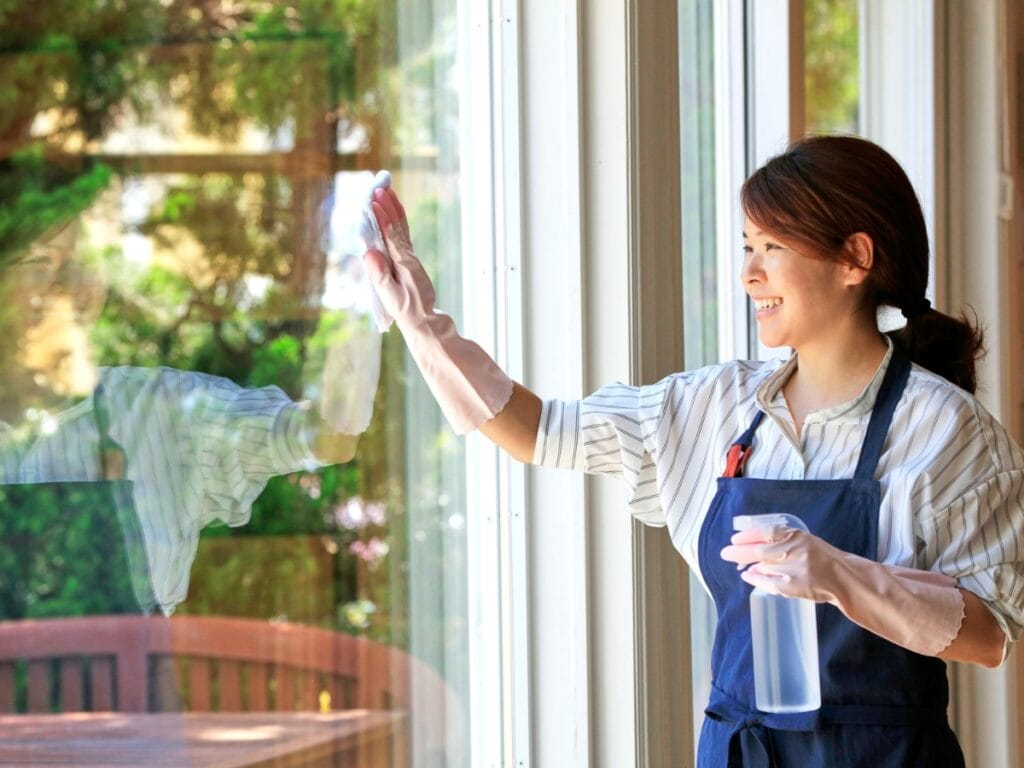 A woman wearing gloves and an apron uses time-saving cleaning hacks as she cleans a large window with a cloth and spray bottle, with trees and outdoor furniture visible outside.