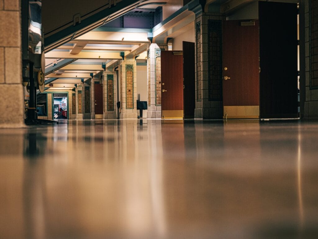 A low-angle view of a brightly lit hallway with closed wooden doors and shiny polished floors, showcasing the results of professional Floor Waxing Services in a public building.