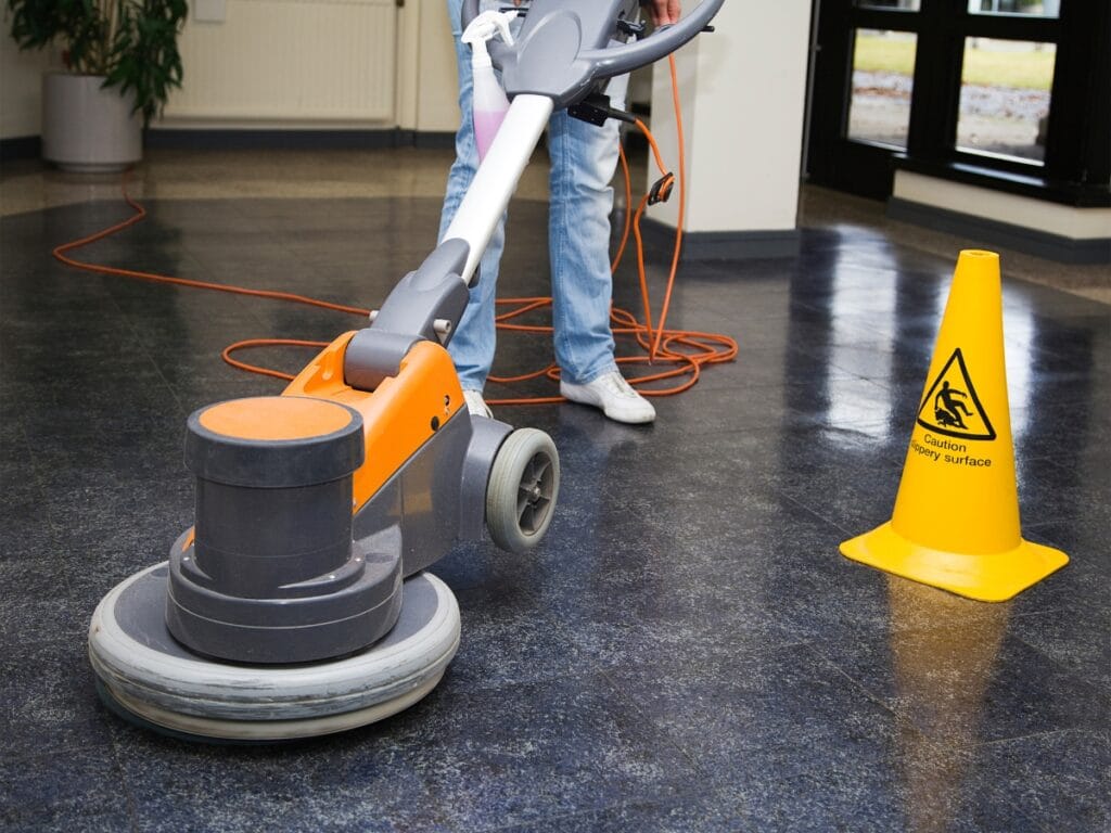A person operates a floor buffer on a shiny dark floor next to a yellow "Caution Wet Floor" sign, with an orange power cord visible&mdash;demonstrating the quality of professional Floor Waxing Services.