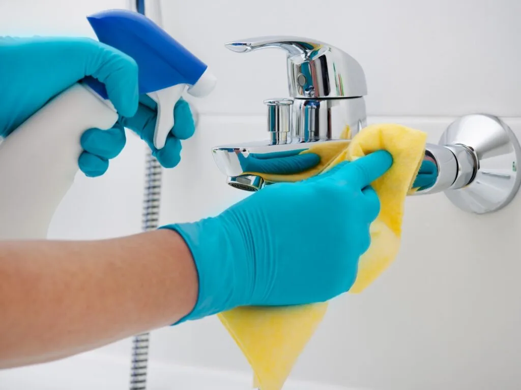 A person wearing blue gloves uses a yellow cloth and cleaning spray to clean a chrome bathroom faucet&mdash;an essential task on any Holiday Cleaning Checklist.