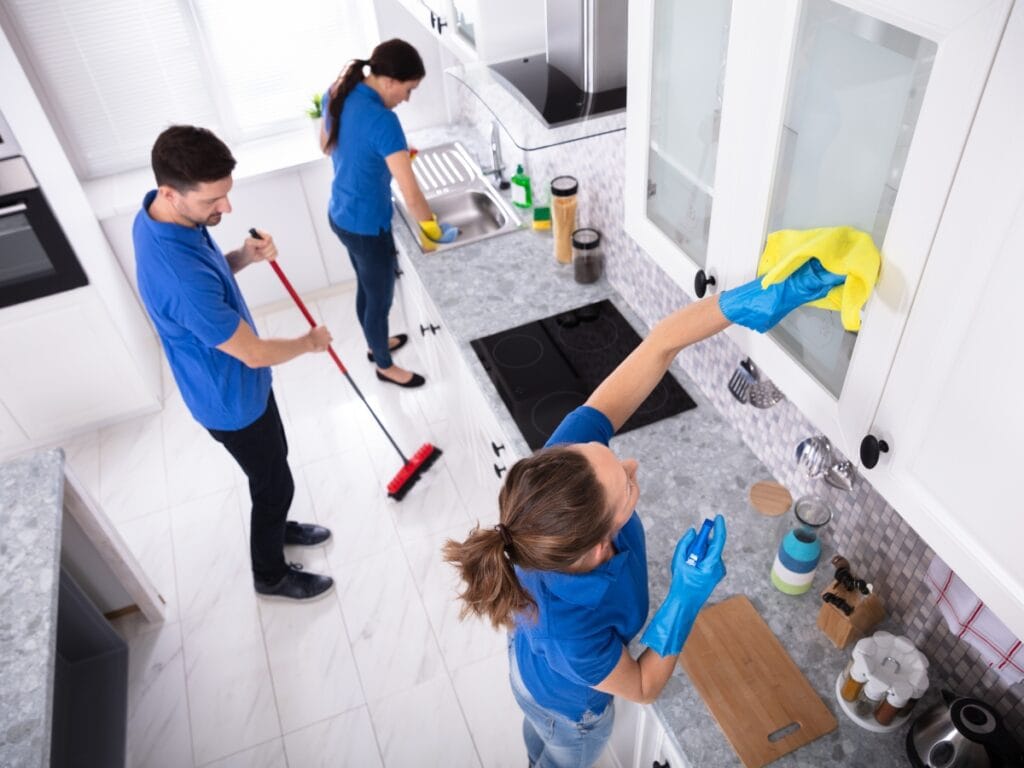 Three people in blue shirts follow a Holiday Cleaning Checklist as they clean a kitchen&mdash;one wipes a cabinet, another sweeps the floor, and the third washes dishes at the sink.