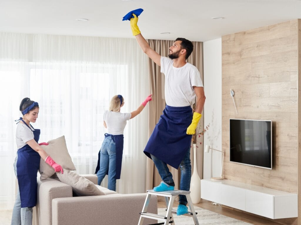 Three people in cleaning uniforms follow a Holiday Cleaning Checklist as they clean a modern living room; one wipes a ceiling fixture from a ladder, while others clean a window and arrange cushions.