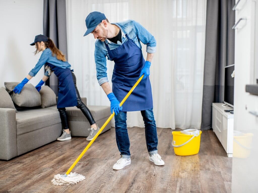 Two people in uniforms and gloves are cleaning a living room; one is mopping the floor, and the other is wiping a sofa. A yellow cleaning bucket sits nearby&mdash;perfect teamwork for any Holiday Cleaning Checklist.