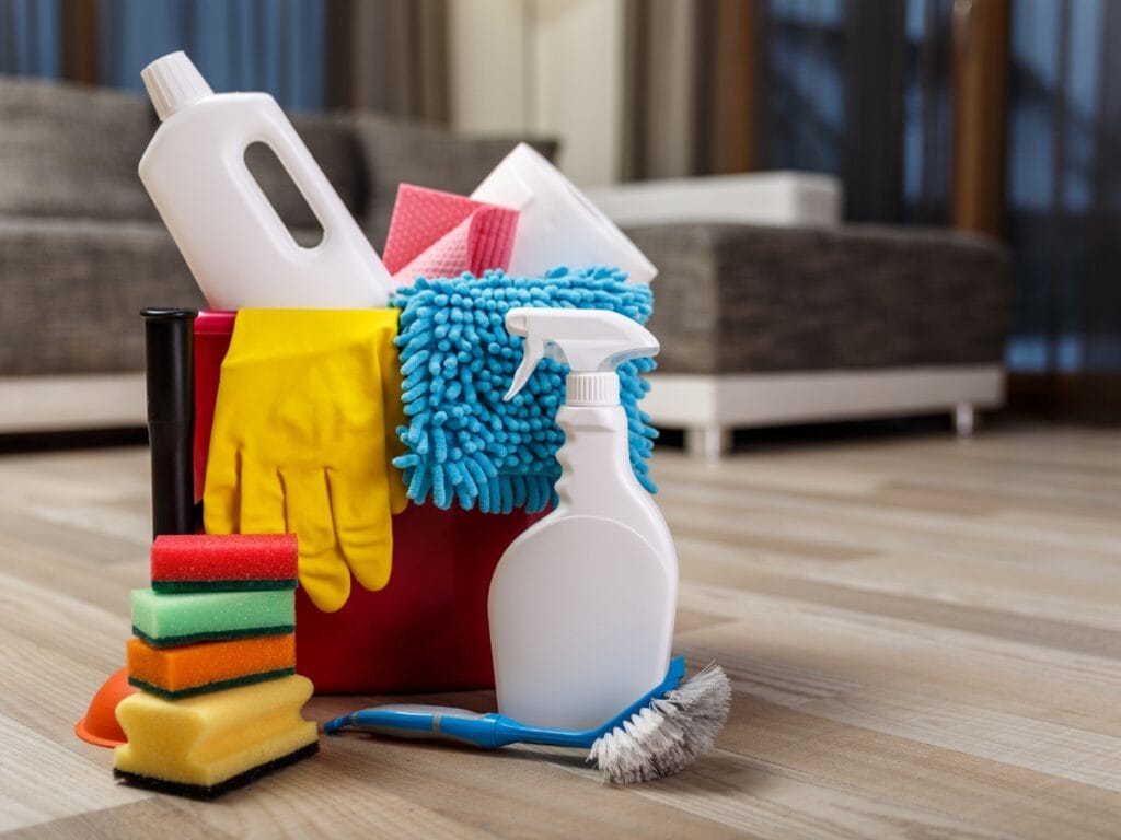 A red bucket filled with cleaning supplies&mdash;including rubber gloves, spray bottle, sponges, brushes, and cloths&mdash;sits on a wooden floor, ready to tackle your Holiday Cleaning Checklist in the living room.