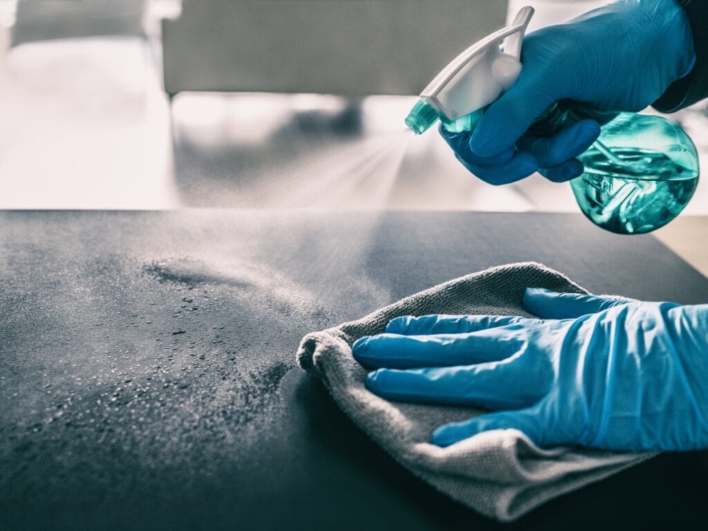 A person wearing blue gloves sprays cleaner onto a surface and wipes it with a cloth, helping to maintain a germ-free home during cold and flu season.