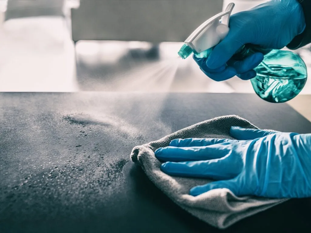A person wearing blue gloves sprays cleaner onto a surface and wipes it with a cloth, helping to maintain a germ-free home during cold and flu season.
