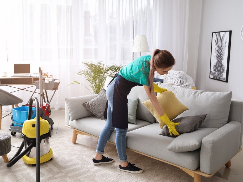 Embracing new year cleaning habits, a woman wearing gloves and an apron arranges cushions on a sofa in a living room, with cleaning supplies and a vacuum cleaner nearby.