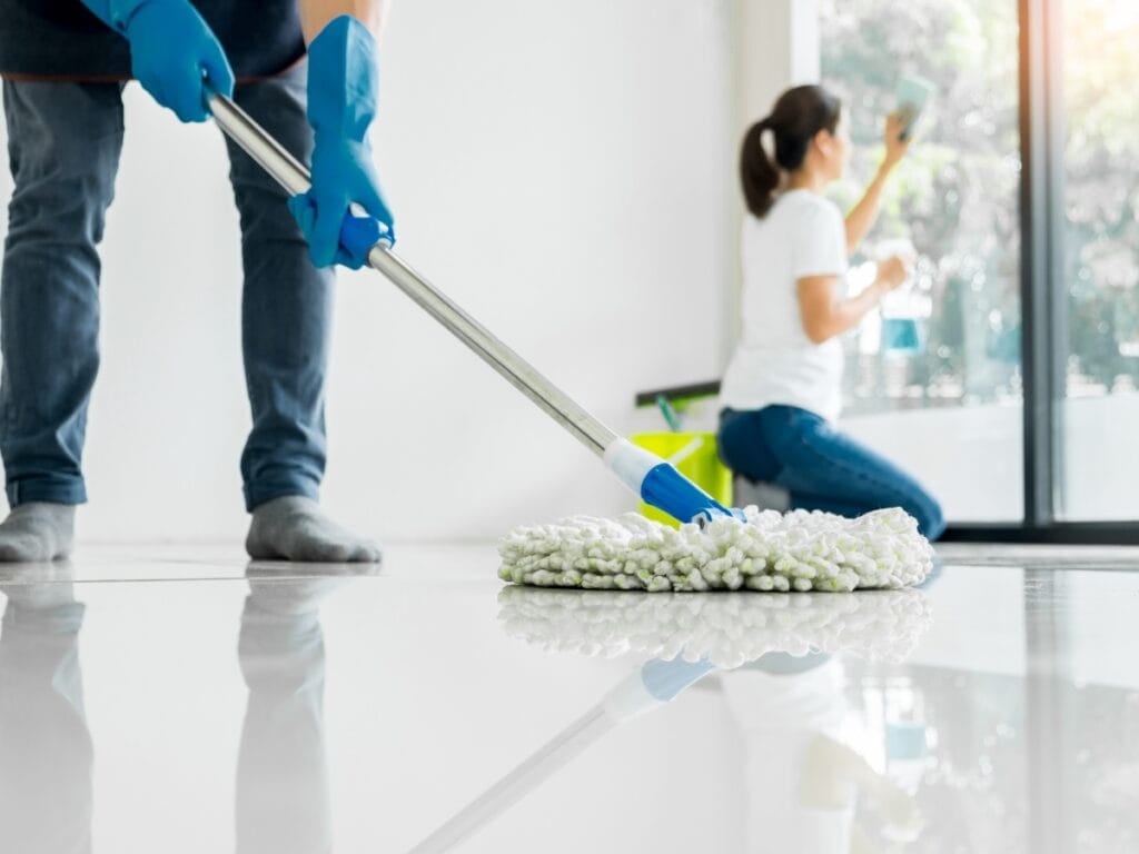 Two people embrace new year cleaning habits in a bright room; one is mopping the floor while another wipes a large window. Cleaning supplies are visible in the background.