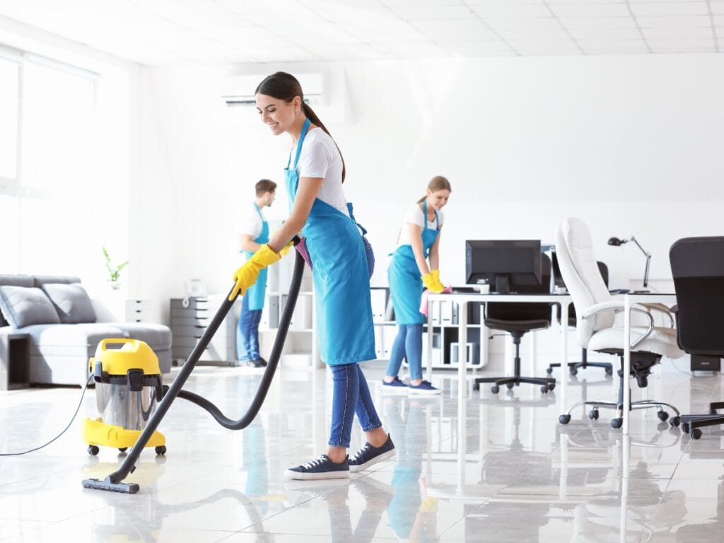 Three people wearing blue aprons and yellow gloves clean a bright, modern office space; offering commercial insights for choosing the best cleaning schedule as one vacuums the floor while two others work at desks in the background.