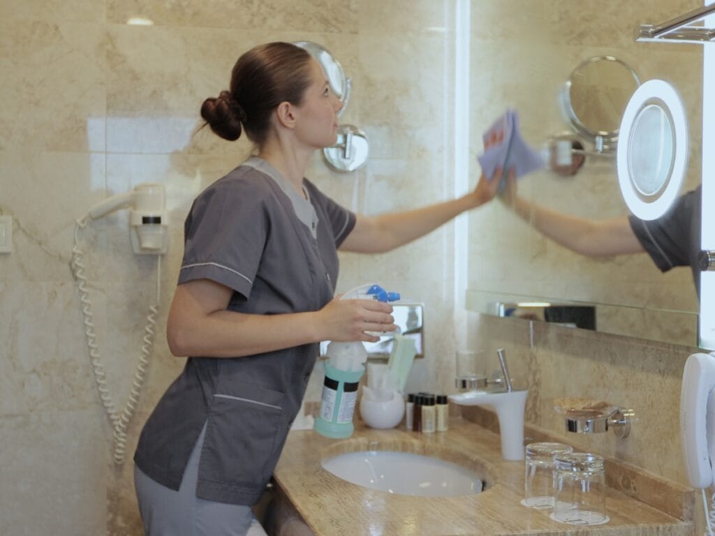 A person in gray uniform cleans a bathroom mirror with a cloth and spray bottle, showcasing specialized services for property management cleaning in Atlanta by the sink and toiletries on a marble countertop.