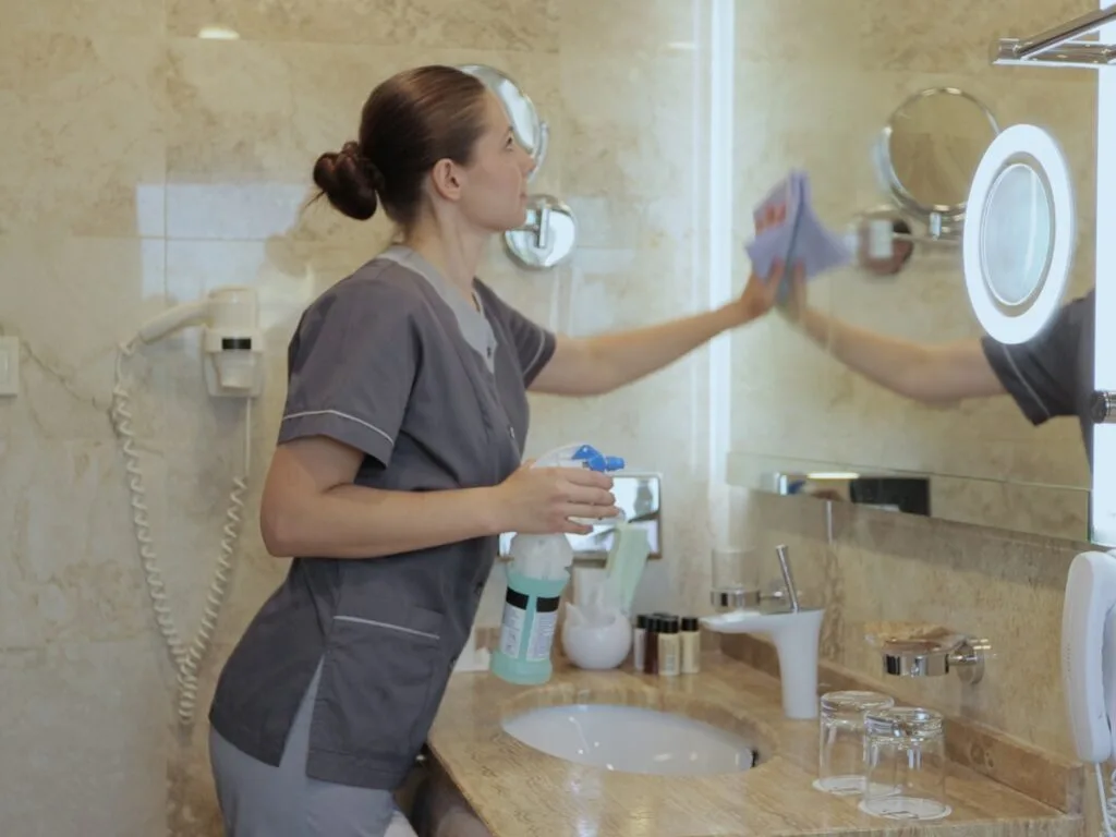 A person in gray uniform cleans a bathroom mirror with a cloth and spray bottle, showcasing specialized services for property management cleaning in Atlanta by the sink and toiletries on a marble countertop.