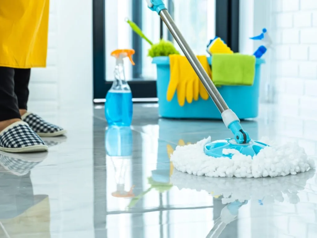 Person mopping a shiny tile floor with a microfiber mop; cleaning supplies, including a blue spray bottle and a bucket, are visible in the background, highlighting the importance of cleanliness in educational settings.