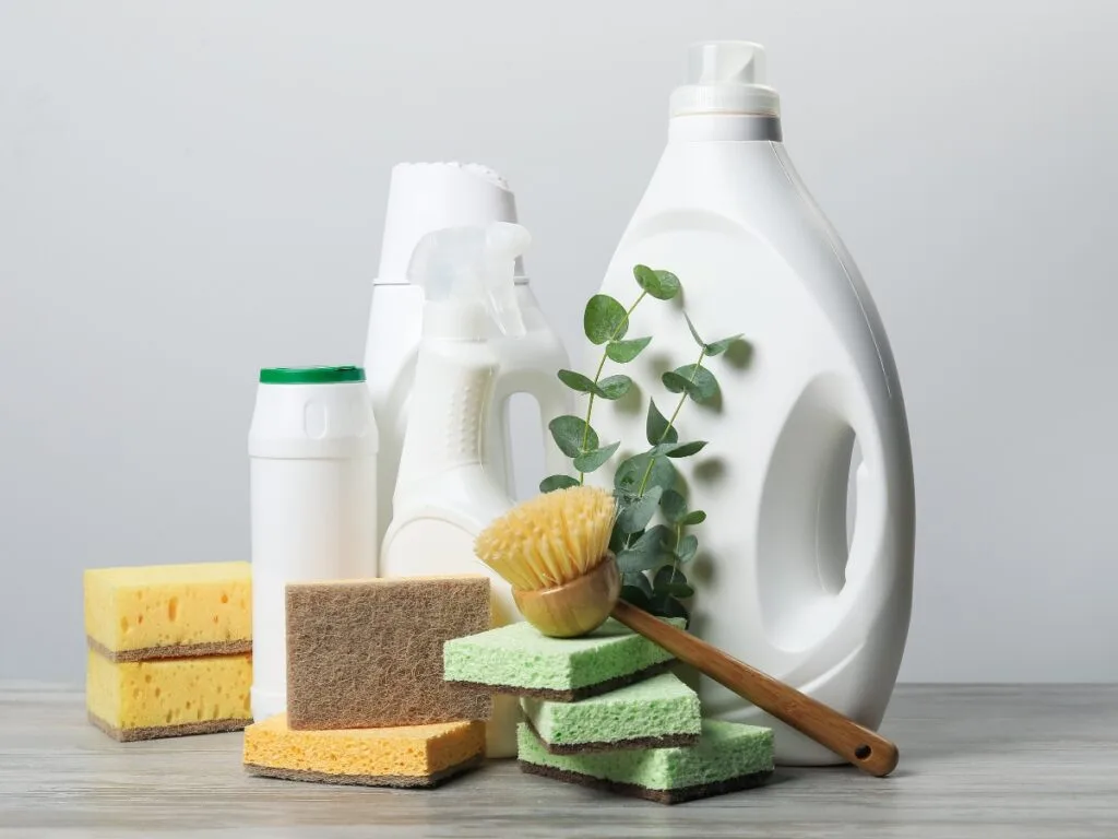 Assorted cleaning supplies, perfect for education on cleanliness, including bottles, sponges, a dish brush, and a sprig of eucalyptus arranged on a wooden surface against a plain background.