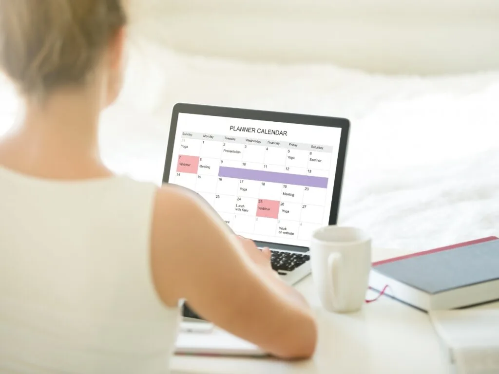 Person sitting at a desk looking at a planner calendar on a laptop, with a notebook and coffee mug nearby—organizing tasks for their education.