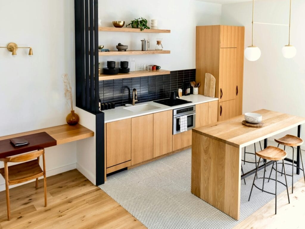 Modern kitchen with stylish wood surfaces, light wood cabinets, black tile backsplash, open shelves, a central island with stools, and a small desk area with a chair on the left.
