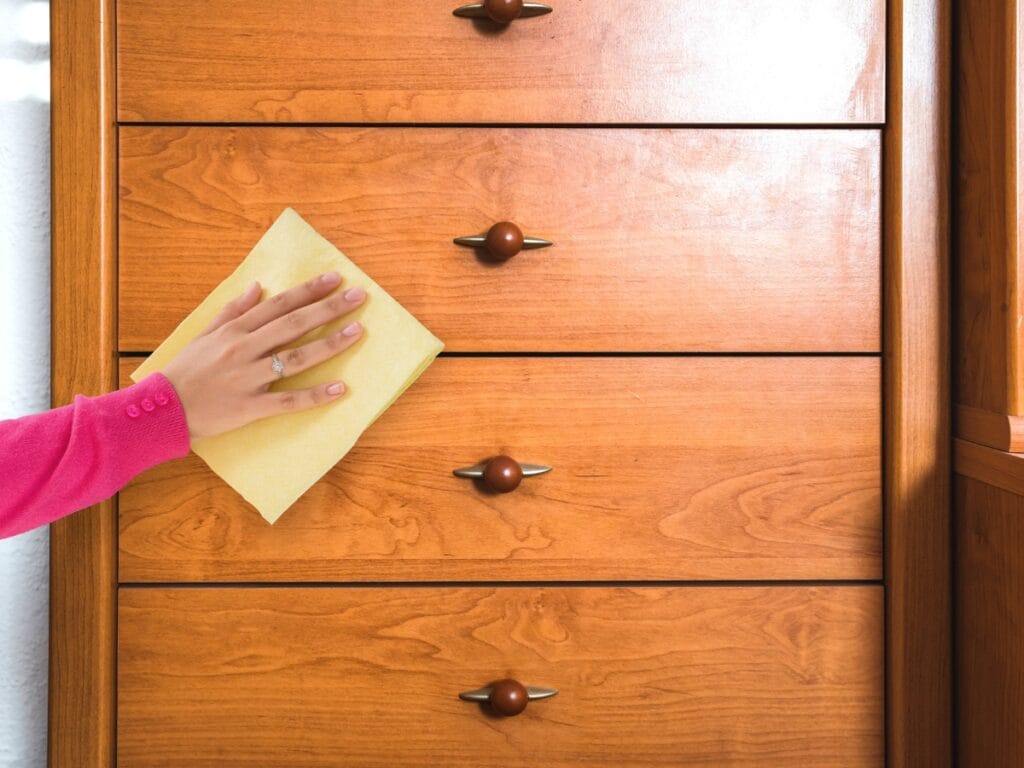 A person in a pink sleeve wipes a wooden dresser with a yellow cloth, gently cleaning the wood surfaces.