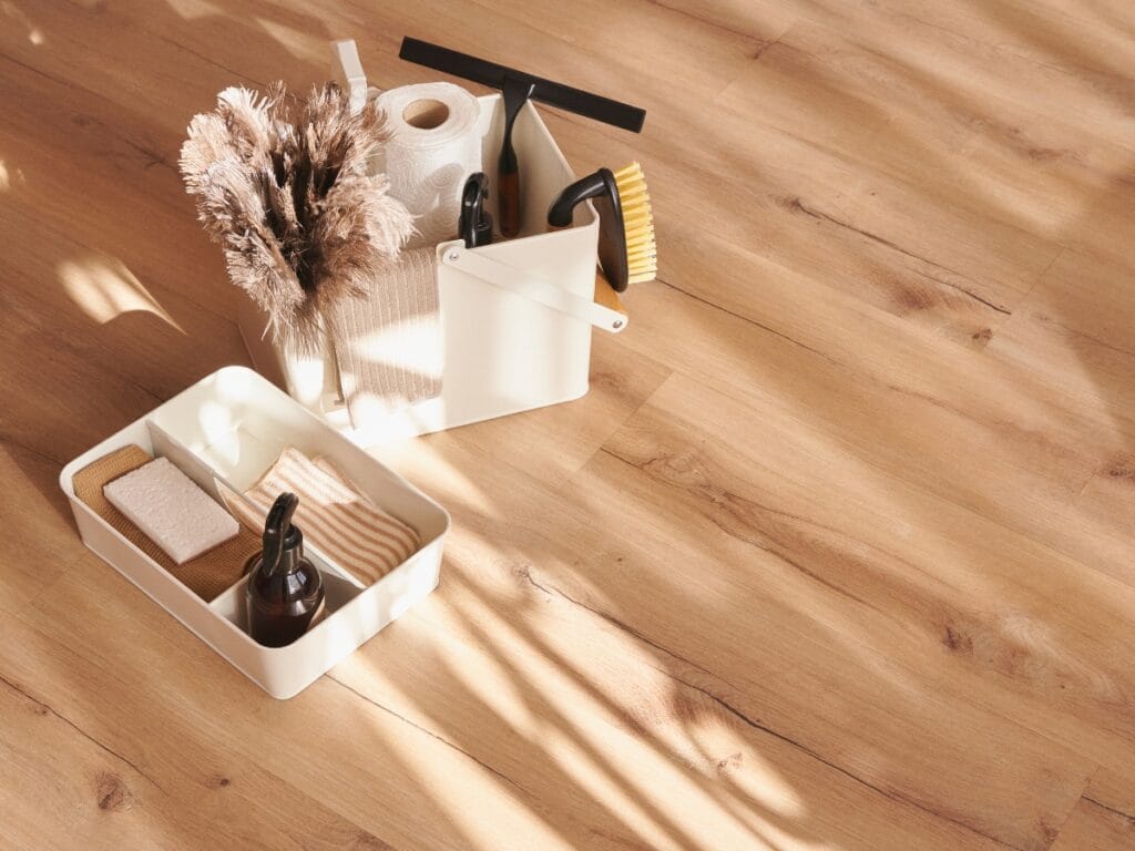 Cleaning supplies, ideal for wood surfaces, including brushes, a spray bottle, sponges, cloths, and a roll of paper towels, are arranged in trays on a light wooden floor with sunlight casting shadows.