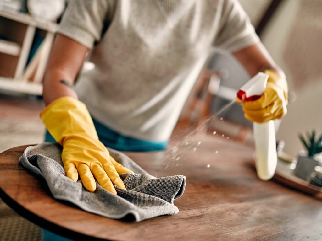 Person wearing yellow rubber gloves sprays cleaner on a cloth while wiping wood surfaces of a table.
