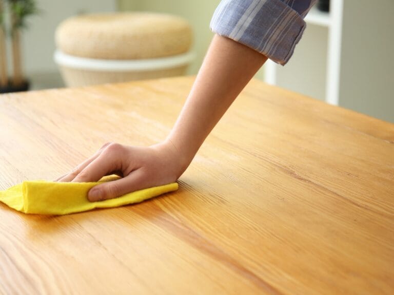 A person wipes a wooden table with a yellow cloth in a well-lit room, taking care to clean and maintain wood surfaces.