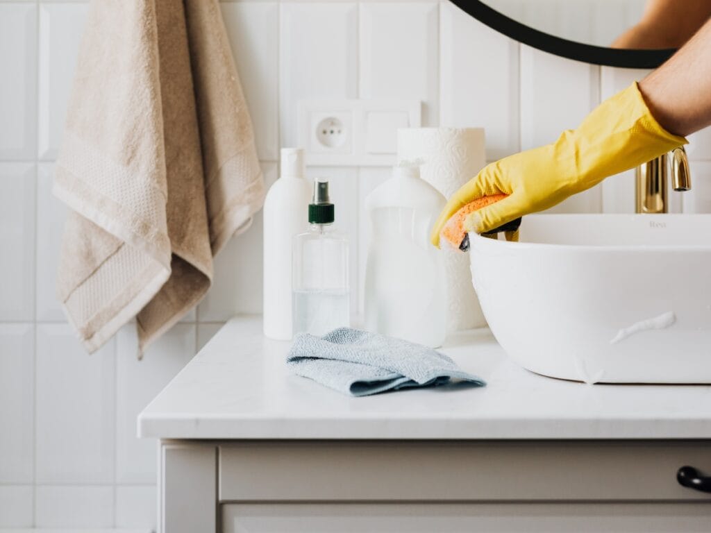 Person wearing a yellow glove cleaning a bathroom sink with a sponge; cleaning products, towel, and toiletries are placed on the countertop for a spotless result—perfect for achieving streak free windows and surfaces throughout your home.